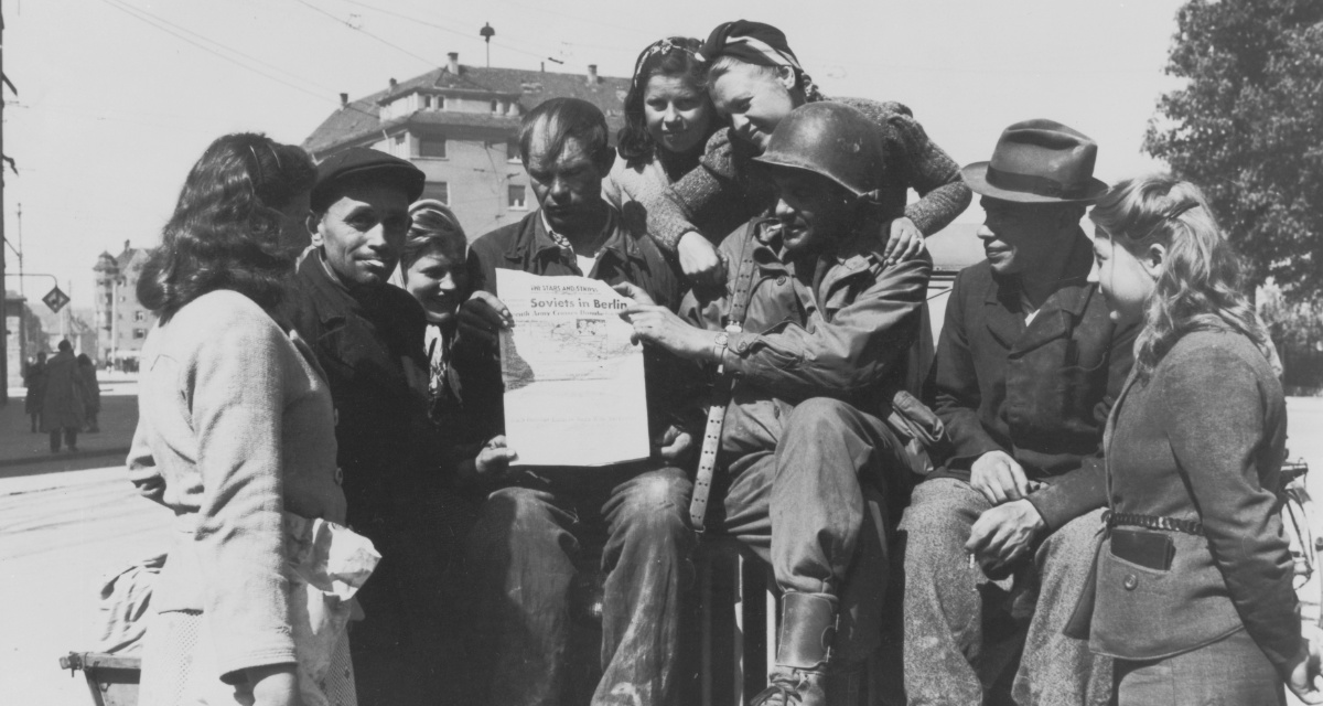 24. April 1945: Ein US-amerikanischer Soldat zeigt sowjetischen Kriegsgefangenen in Ulm eine Zeitung, in der über die Befreiung Berlins durch sowjetische Truppen berichtet wird.